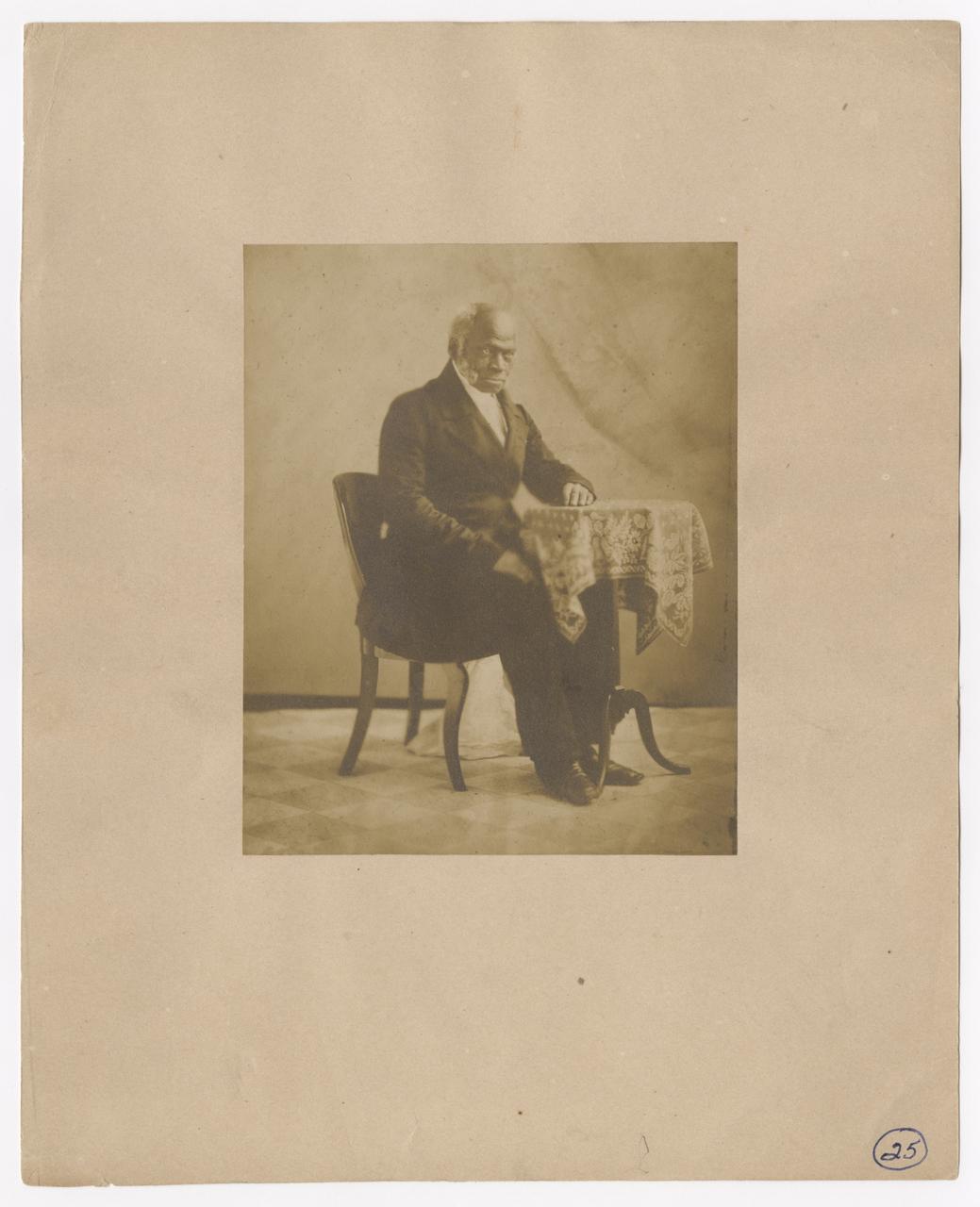 Toussaint sits on a chair with his left arm resting on a side table.