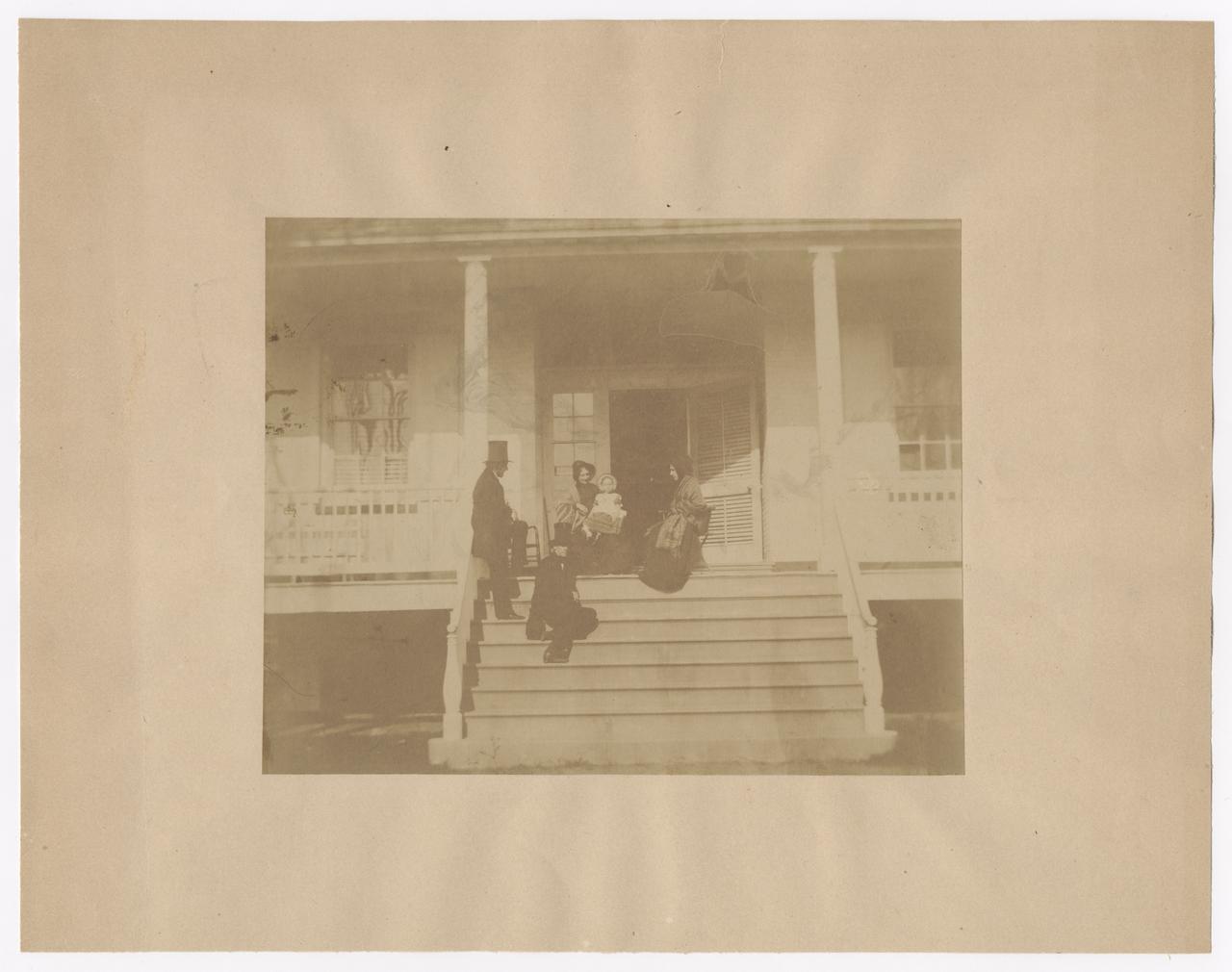 Two men and two women sitting on a porch. One of the women is holding on a young child.