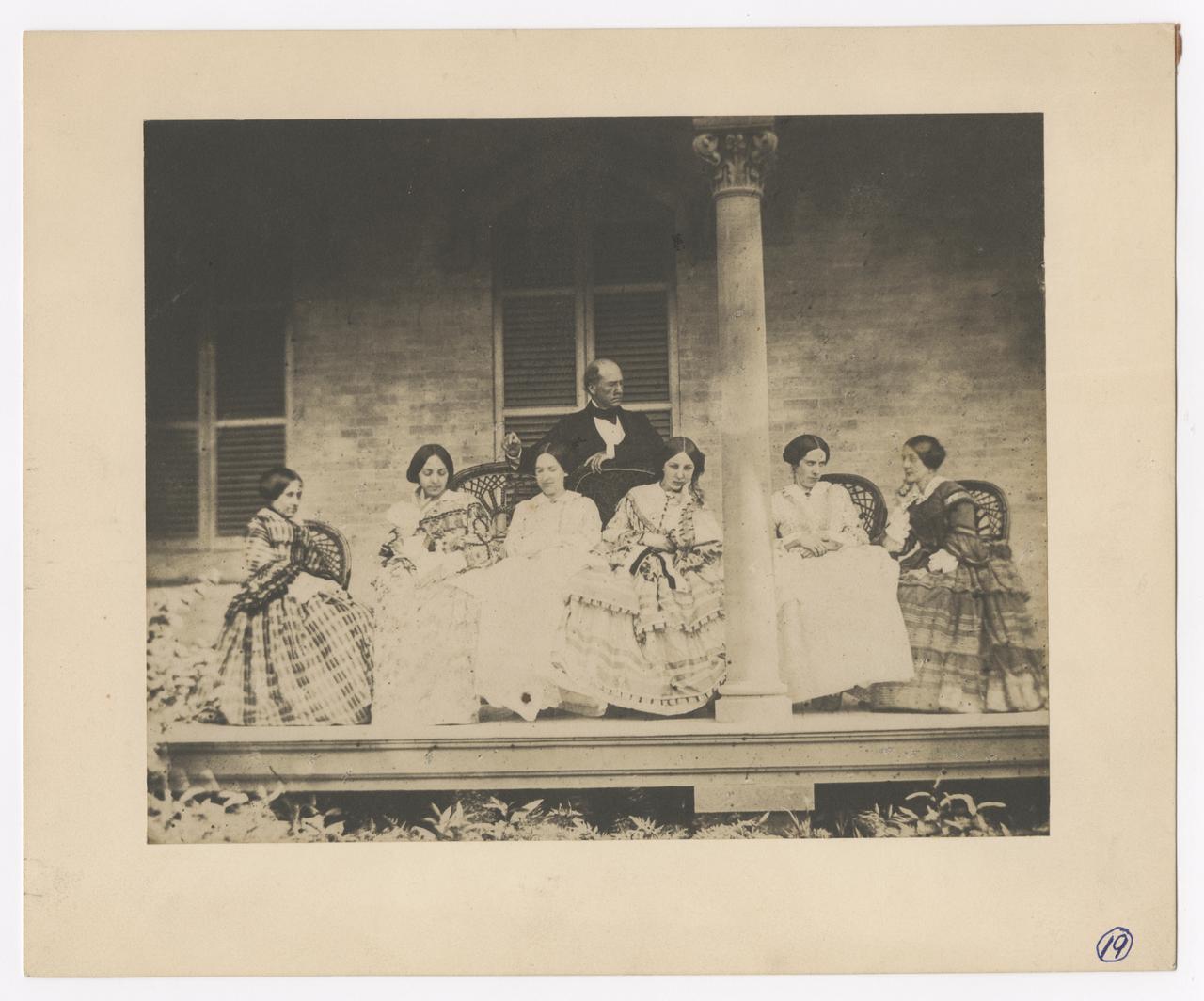 Group of seven on a porch. Six women seated, with one man standing behind them.