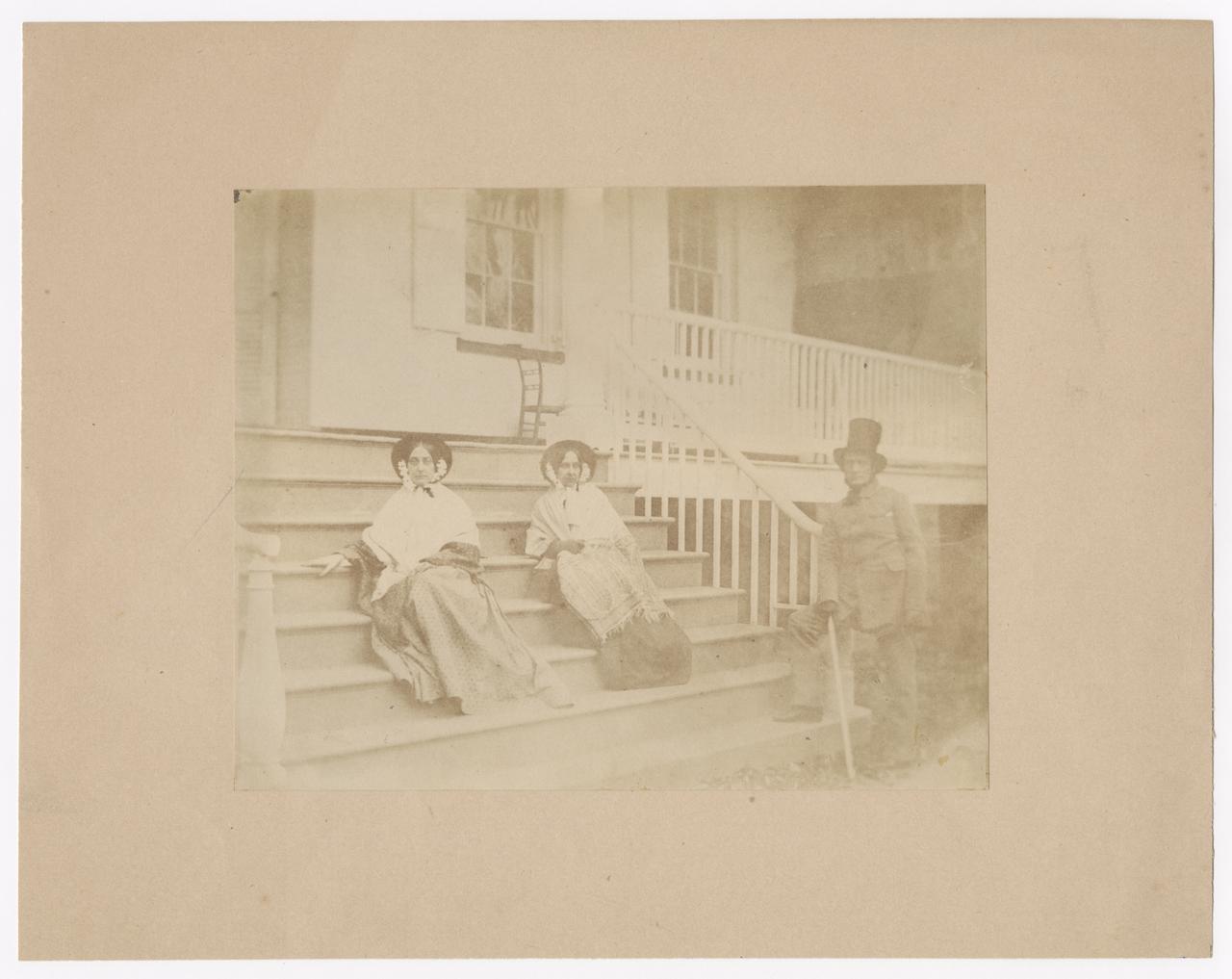 Two women seated and one man standing on the porch steps.