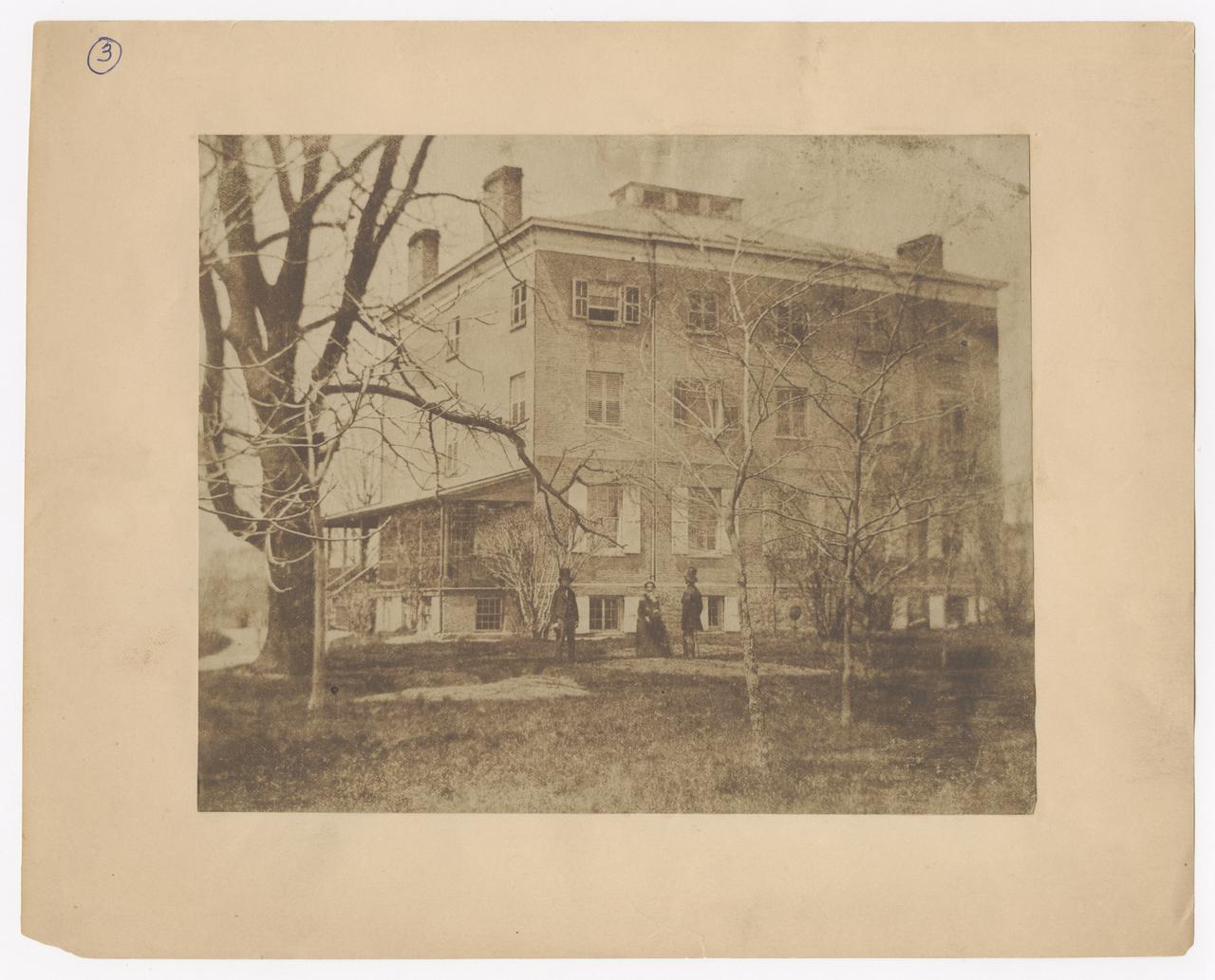 View of the exterior of Chelsea house and the surrounding trees. Two men and a woman stand in front of the house.