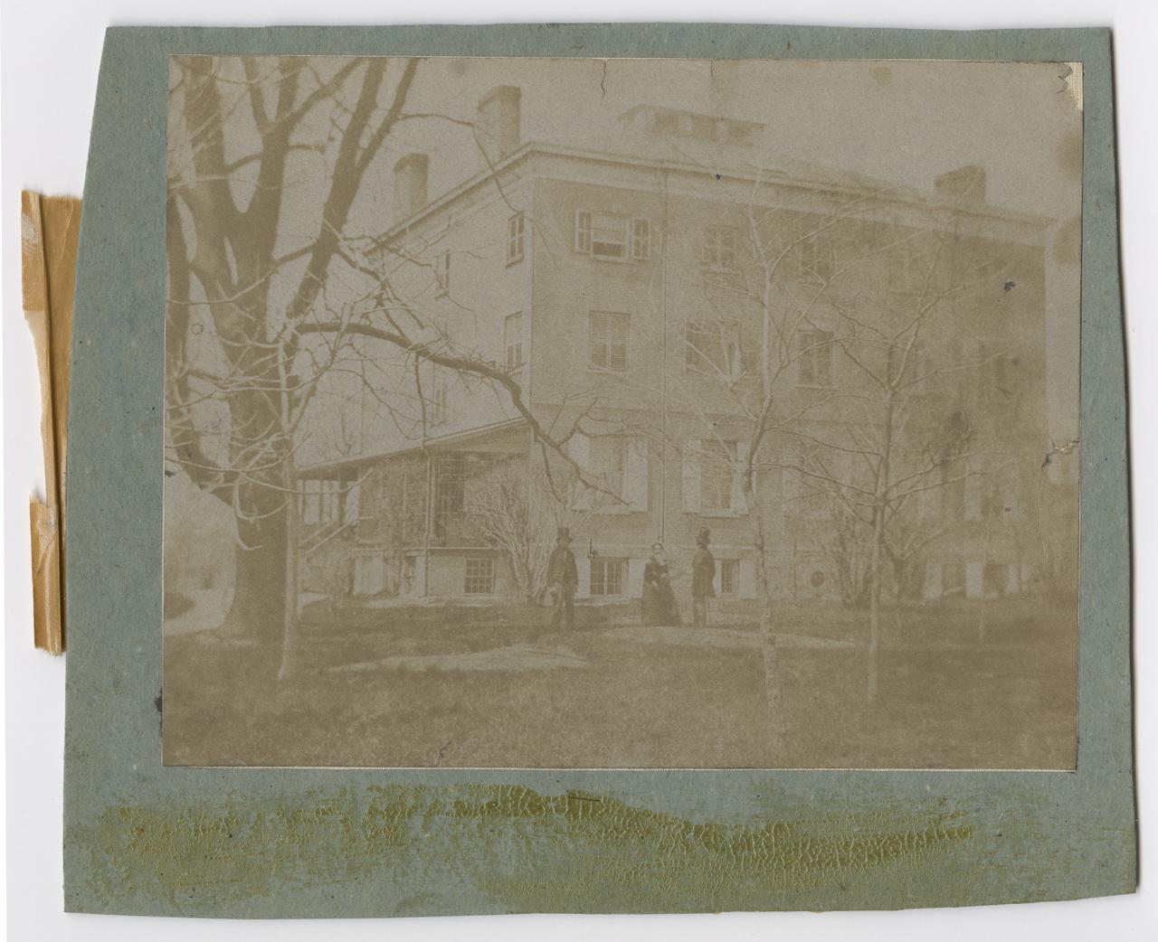 View of the exterior of Chelsea house and the surrounding trees. Two men and a woman stand in front of the house.