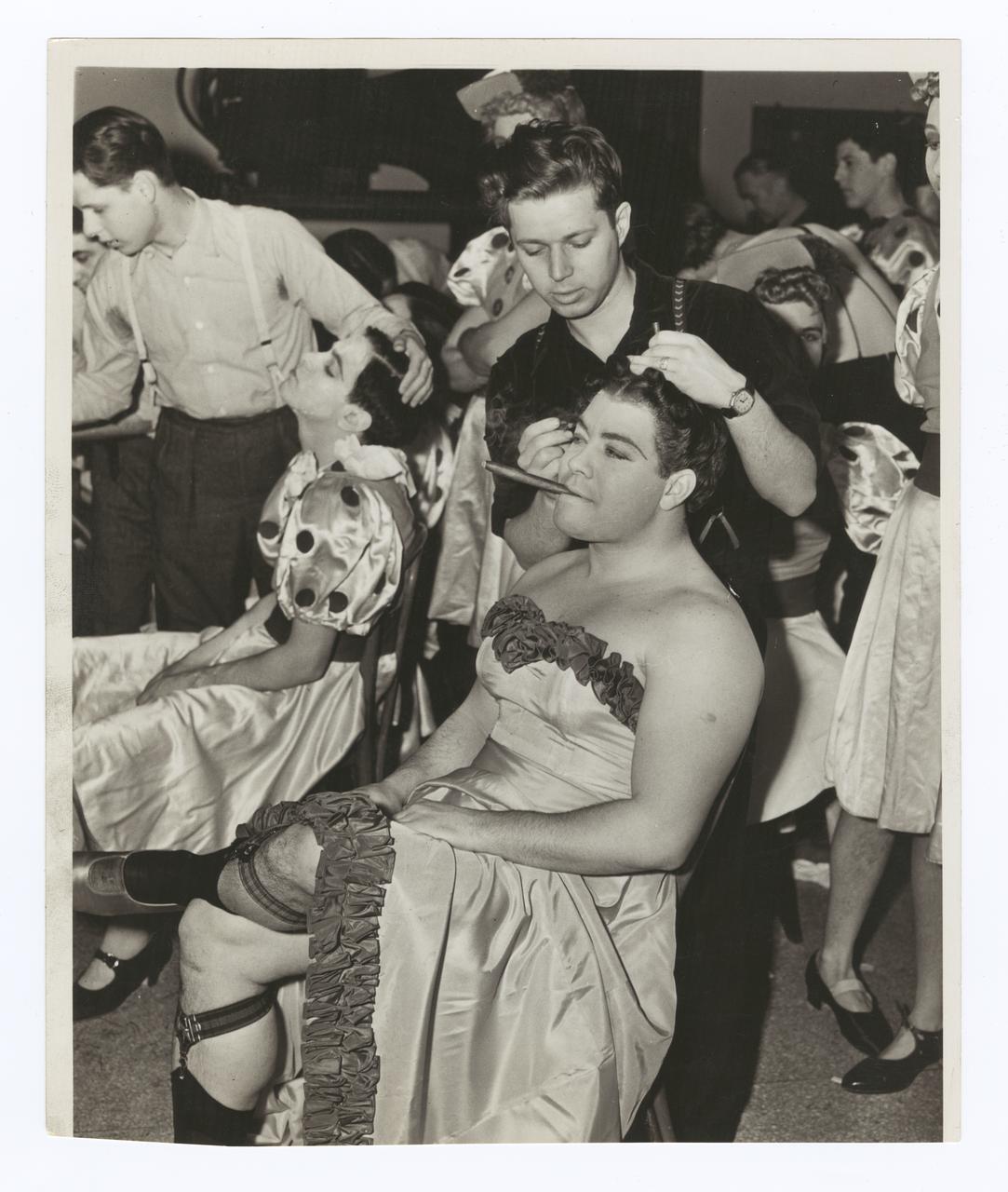 John Bateman getting his hair and makeup done for 1939 Varsity Show Fair Enough. He is sitting in a chair smoking a cigar and wearing a dress.